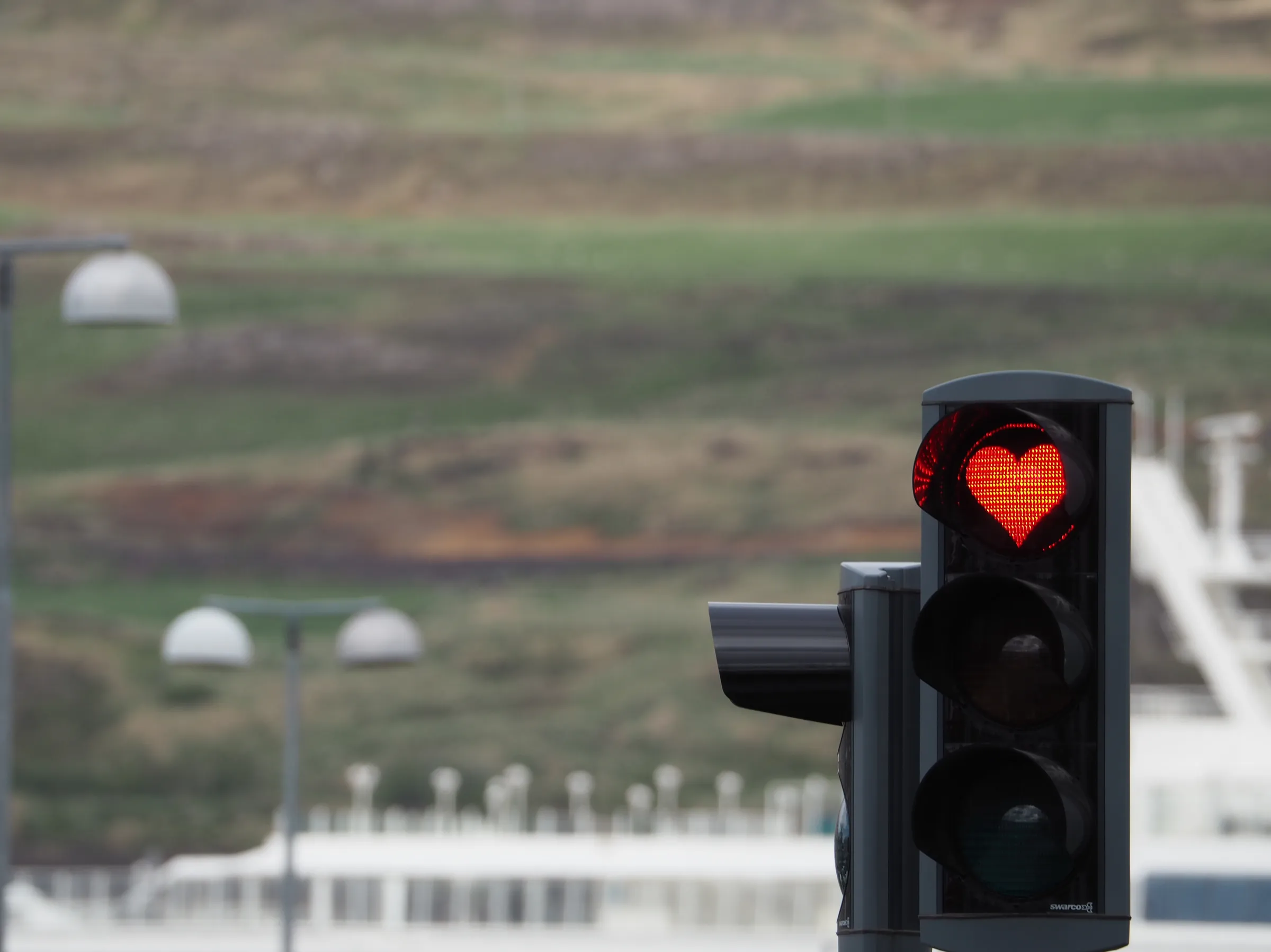 A traffic light in Akureyri glowing with a red heart, with a cruise ship and the hillside across the fjord behind it